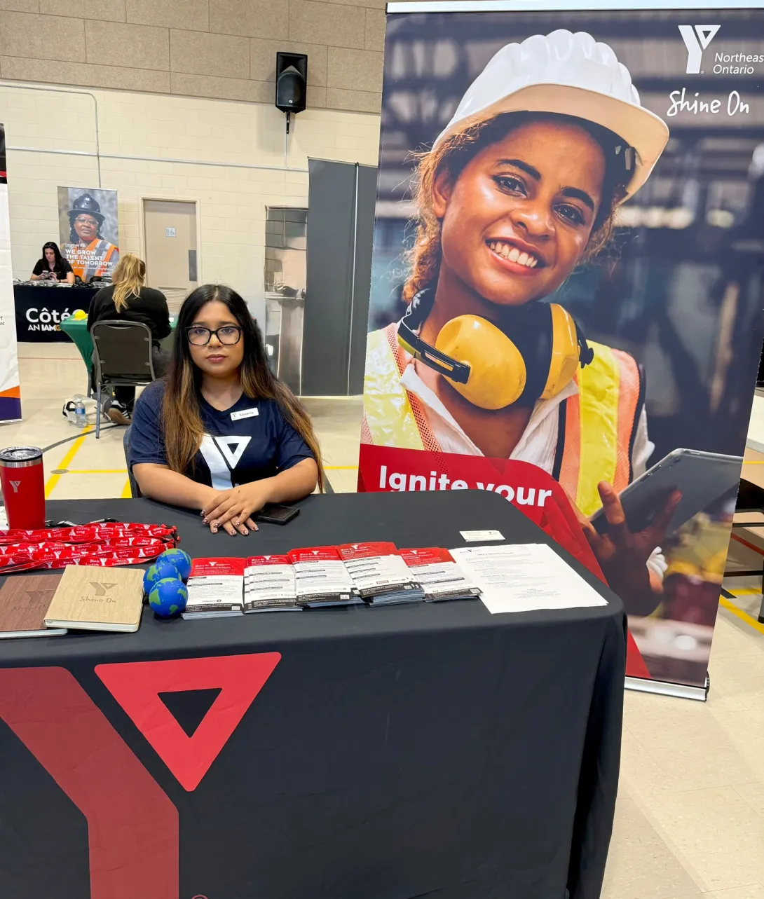 Samanta Saif Kristy sitting behind a YMCA promotional table lined with brochures and informational materials, next to a large "Shine On" YMCA banner.