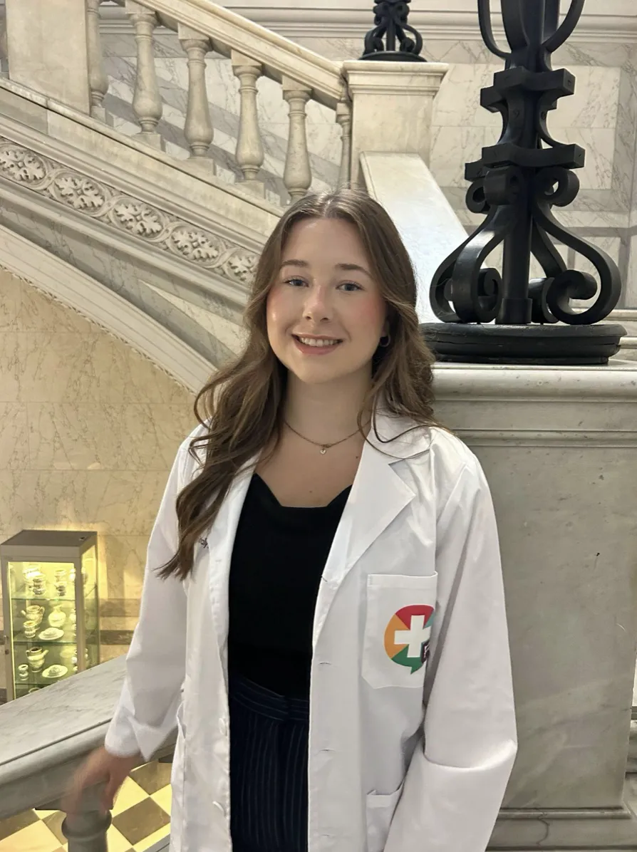 Kaleigh Hamilton smiling while wearing a white lab coat over a black top, standing indoors next to a marble staircase with decorative iron balustrades.