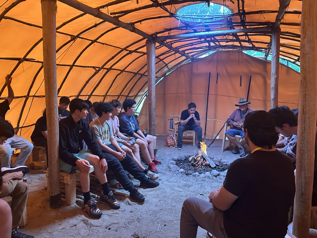 A group of OYEP participants and leaders sitting in a circle inside a traditional teaching lodge around a burning sacred fire.
