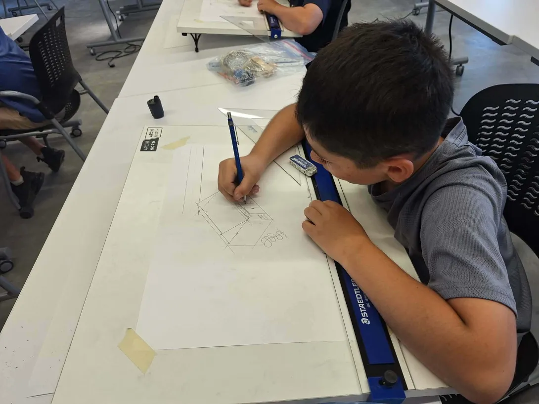 A young student leans over a drafting board, using a pencil and ruler to carefully draw a 3D architectural sketch of a house during the Archi·North Summer Camp at Laurentian University.