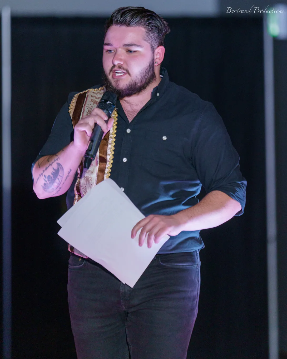 Connor Lafortune holding notes and speaking into a microphone on stage, wearing a black shirt with traditional ribbons.
