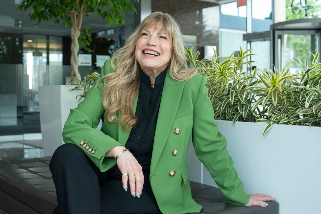 Candid portrait of Rachel Prudhomme laughing while leaning back against a bench in a bright modern atrium, dressed in a vibrant green double-breasted blazer over a black top and trousers, surrounded by indoor green plants.