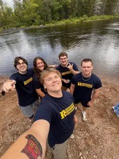 A group of five smiling Laurentian University Residence Assistants (RAs) pose outdoors on a dirt shore in front of a wide river lined with lush green trees. Grant Petronech holds the camera in the foreground taking a selfie, wearing a dark blue t-shirt with yellow Laurentian Residence lettering and showing a red tattoo on his inner forearm. Four other RAs stand close together behind him, all dressed in matching navy Laurentian shirts, smiling and making casual hand gestures toward the camera.
