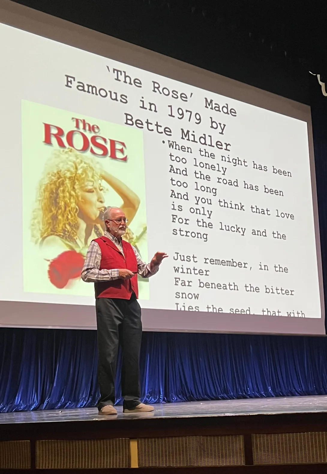 Dr. Joe Shorthouse speaking on stage in front of a presentation slide featuring Bette Midler's album cover for "The Rose" alongside song lyrics. He is wearing a red vest over a plaid shirt and dark trousers while pointing toward the slide.