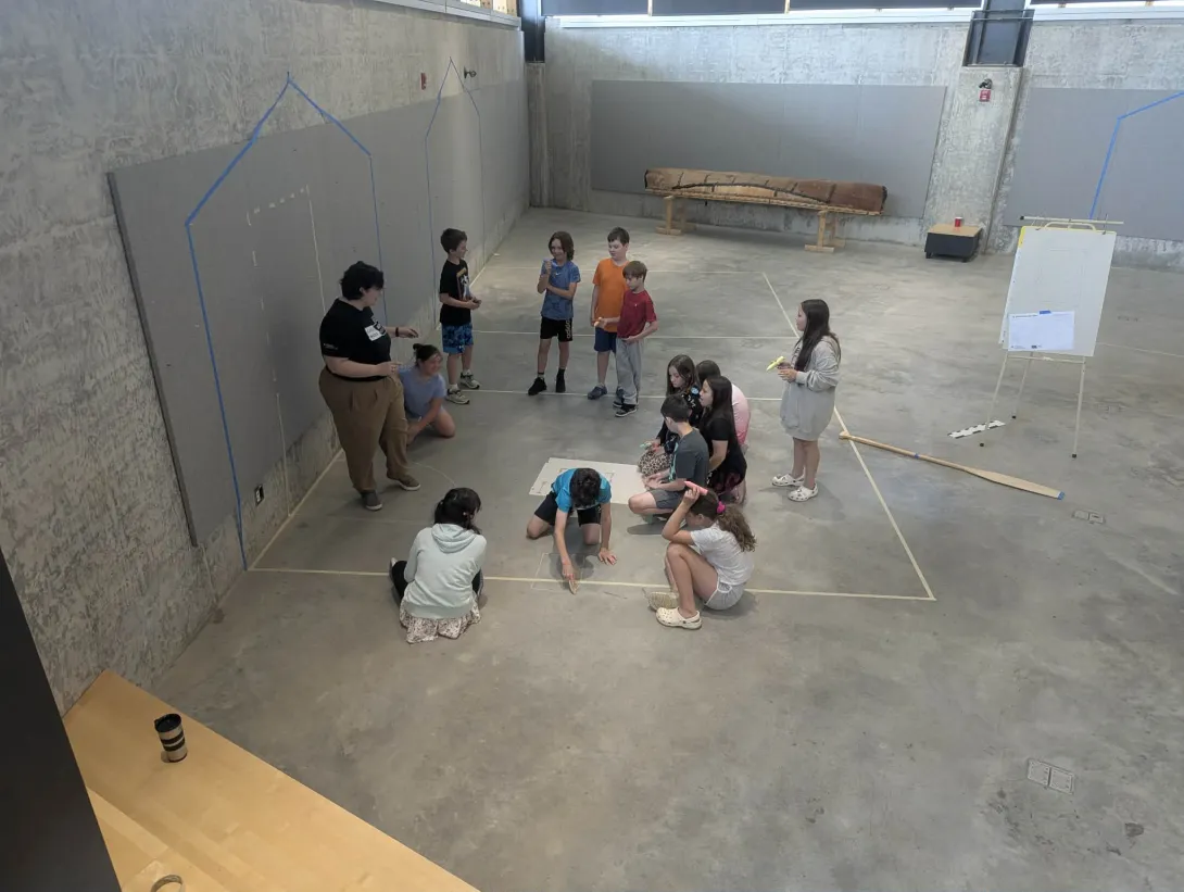 An instructor guides a group of campers in a studio space as they collaborate on floor plan layouts taped directly onto the floor.
