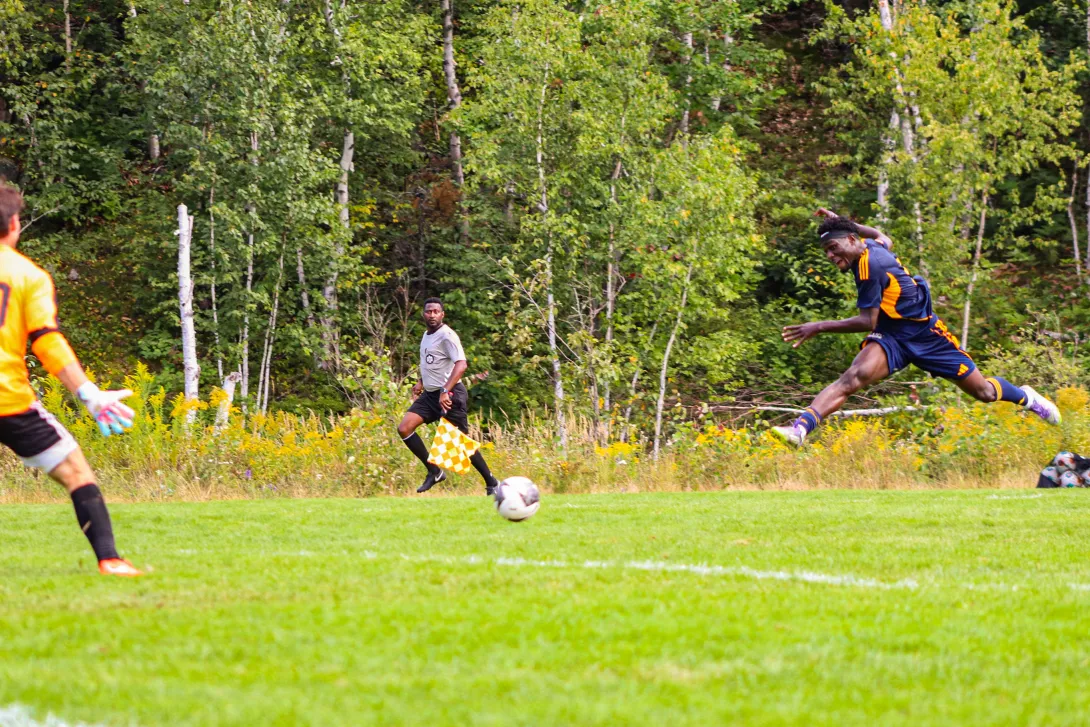 A Laurentian Voyageurs men's soccer player leaping through the air to take a shot on goal during an outdoor match, with an opposing goalkeeper in orange attempting to block and a line referee visible in the background.