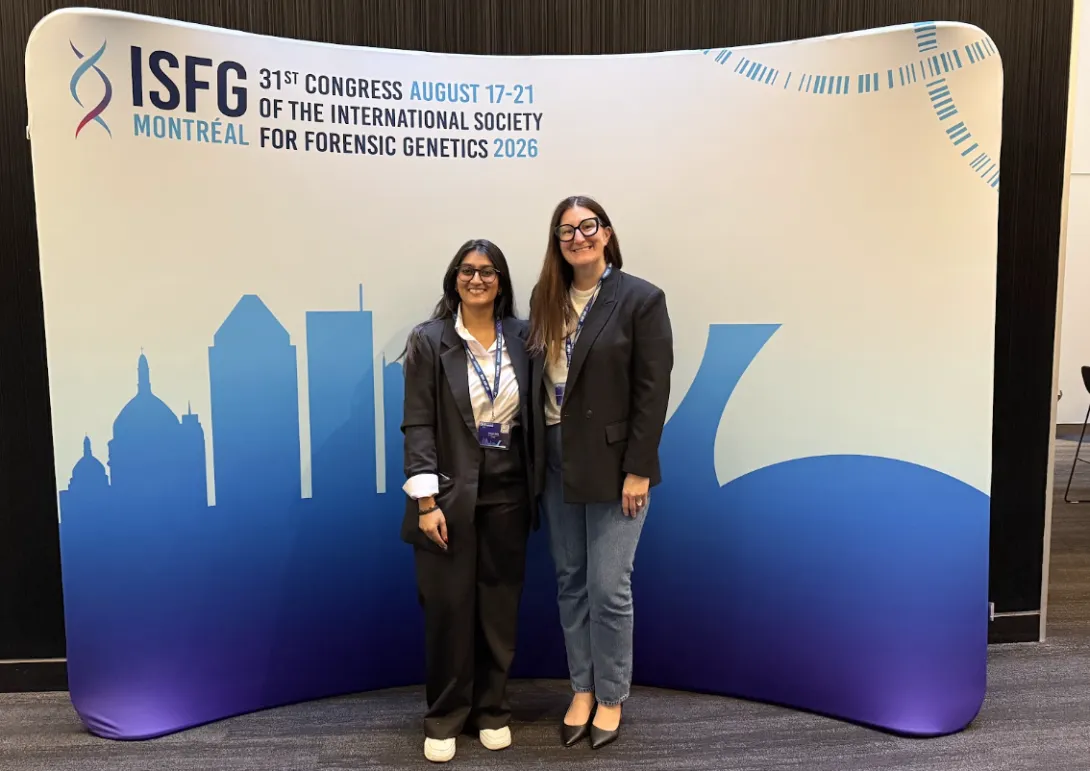 Shivani Gami standing alongside her co-supervisor, Dr. Nicole Novroski, in front of a white and blue ISFG 2026 backdrop featuring the Montreal skyline and DNA strand graphics. Both are dressed in professional attire and wearing conference lanyards.