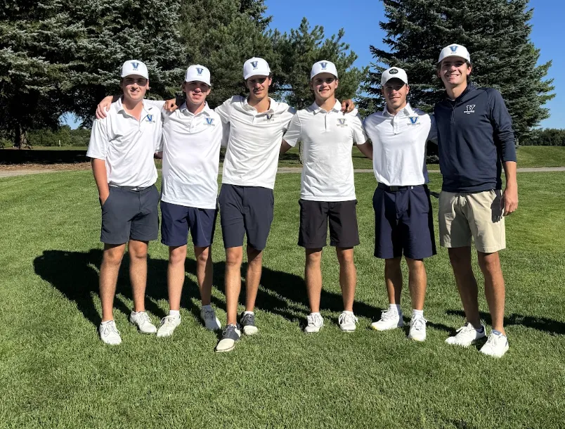 Six male golfers from the Laurentian Voyageurs team stand arm-in-arm on a sunny green golf course framed by tall evergreen trees. Five members wear white polo shirts, dark shorts, and white athletic caps with the team logo, while the person on the far right wears a dark blue long-sleeve athletic pullover.