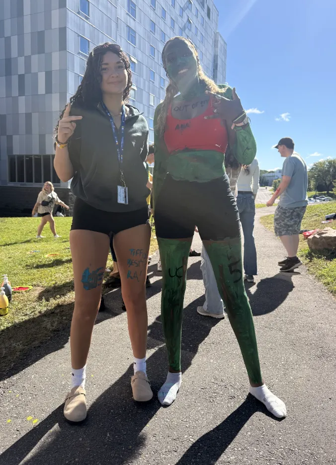 Residence Assistant Tiffany Uzamere covered in green body paint over a red top and black shorts, standing outdoors on a paved walkway next to another smiling RA near a multi-story campus residence building on a sunny day.
