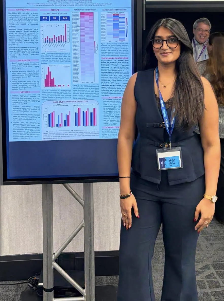 Shivani Gami standing beside her forensic genetics research poster at the ISFG Congress in Montreal, wearing a dark blue vest and matching trousers. In the background over her left shoulder, world-renowned forensic geneticist Dr. John Butler smiles directly at the camera, photobombing the shot.