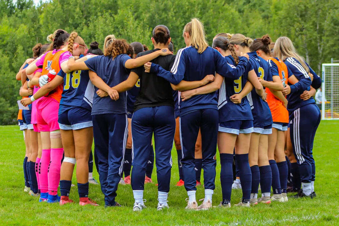 The Laurentian Voyageurs women's soccer team seen from behind, gathered in a large team huddle on a grass field with their arms around each other, wearing blue, pink, and orange athletic apparel.