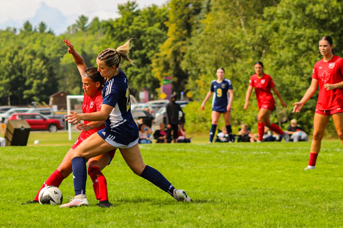 A high-action photo of a Laurentian Voyageurs women's soccer player in a dark blue kit battling an opposing player in red for possession of the ball on an outdoor grass pitch, with teammates and spectators watching in the background.