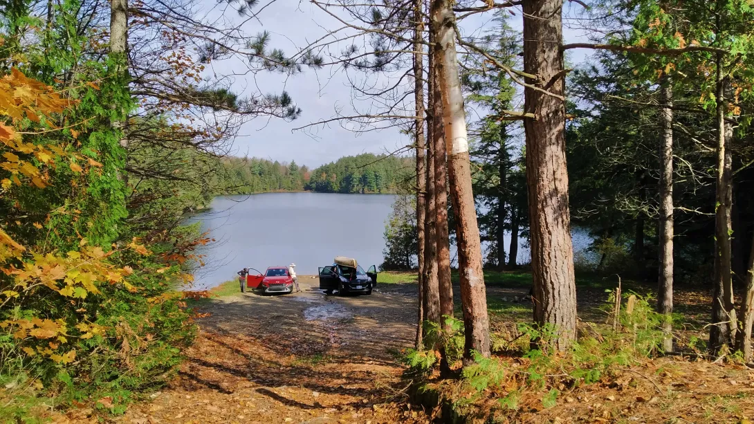 A scenic view framed by tall forest trees looking down toward a tranquil lake landing, where two vehicles are parked on the shore surrounded by autumn foliage.