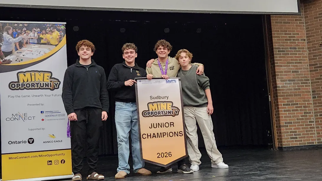 A team of four high school boys stand together on stage smiling while holding the "Sudbury MINE OPPORTUNITY JUNIOR CHAMPIONS 2026" award banner.